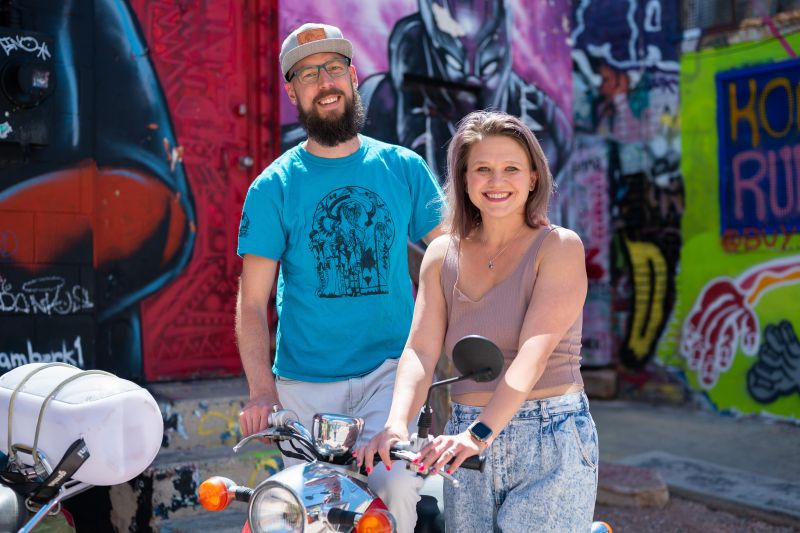 Michael and Jennifer Calabrese with moped in art alley downtown rapid city. mural of black panther behind couple. he had blue shirt and grey baseball cap. Jennifer wears jeans with light purple tank top.